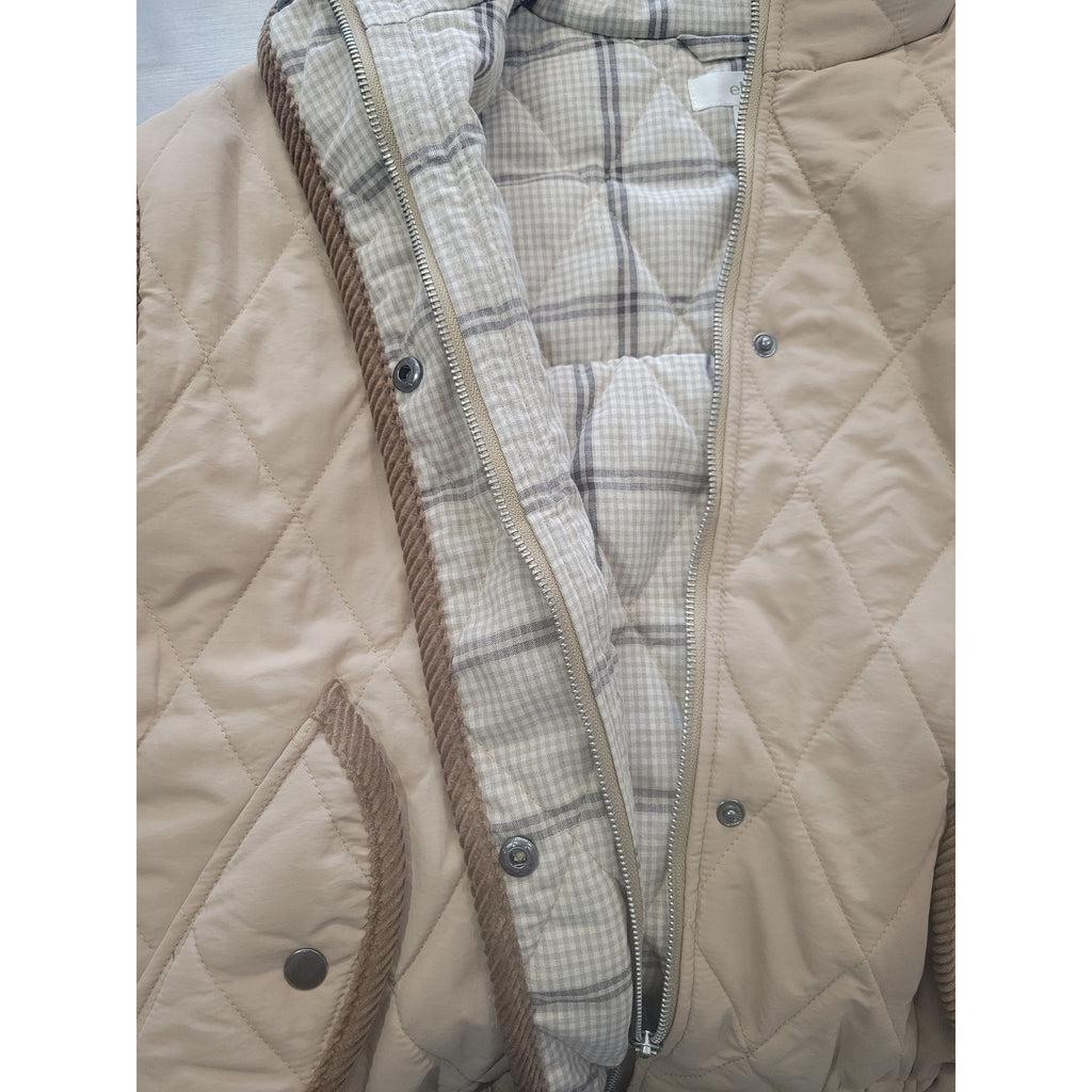 Close-up of a quilted beige jacket or quilted blanket with a diagonal zipper opening, showing a plaid patterned lining and brown cord trim.